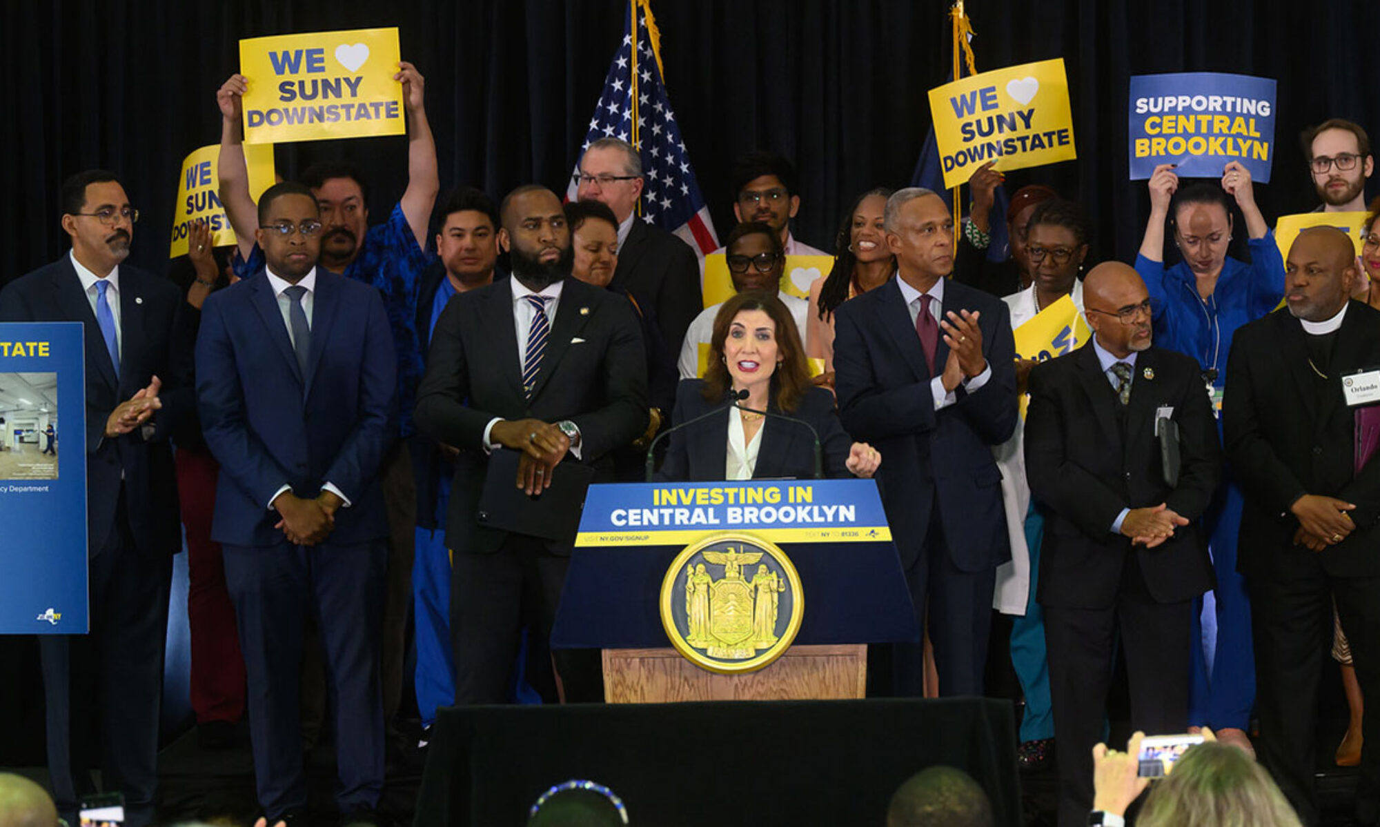Governor Hochul stands behind a posium on stage with many state leaders behind her during a press event announcing updates to SUNY Downstate Medical Center.